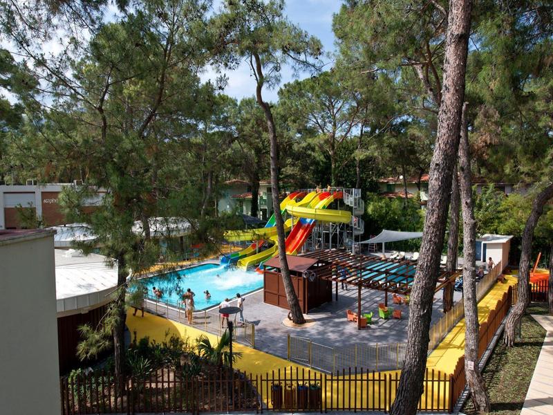 Freizeitpark mit Rutschen, Pool, Bäumen und Holzelementen, umgeben von einem Zaun.