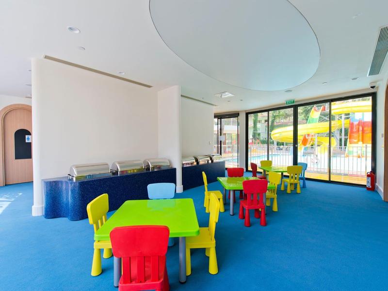 Heller Raum mit bunten Kindertischen und Stühlen auf blauem Boden, großer Glasfront und weißer Decke.