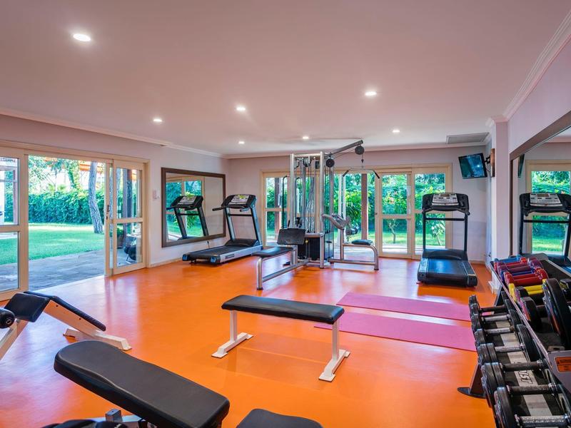 Helles Fitnessstudio mit orangem Boden, verschiedenen Trainingsgeräten und großen Fenstern zum Garten.