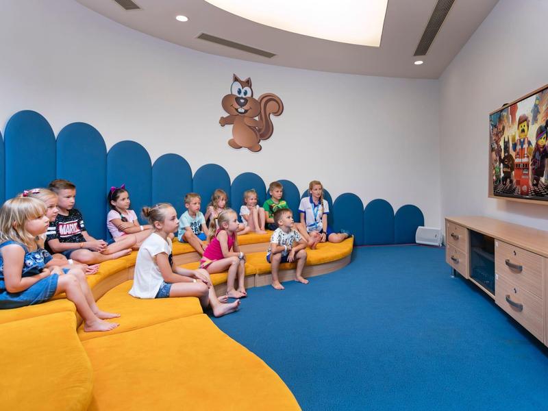 Kinder sitzen in einem bunten Raum auf gelben Sitzbänken und schauen gespannt einen Film.