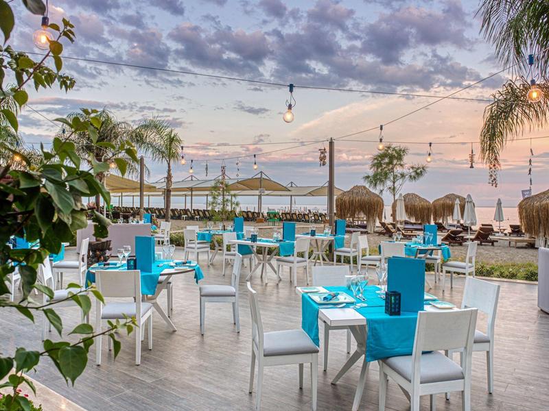 Strandrestaurant mit weißen Stühlen, blauen Tischdecken, Palmen und Wolkenhimmel bei Sonnenuntergang.