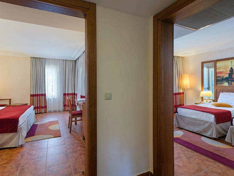 Innenansicht mit zwei angrenzenden Hotelzimmern, je mit Bett, Stuhl, Tisch und warmem Licht.