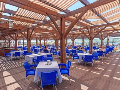 Duży zadaszony taras z niebieskimi krzesłami i białymi stołami w słonecznym otoczeniu.