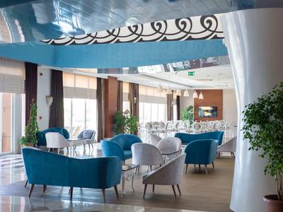 Jasny salon hotelowy z niebieskimi i szarymi fotelami oraz roślinami przed dużymi oknami.