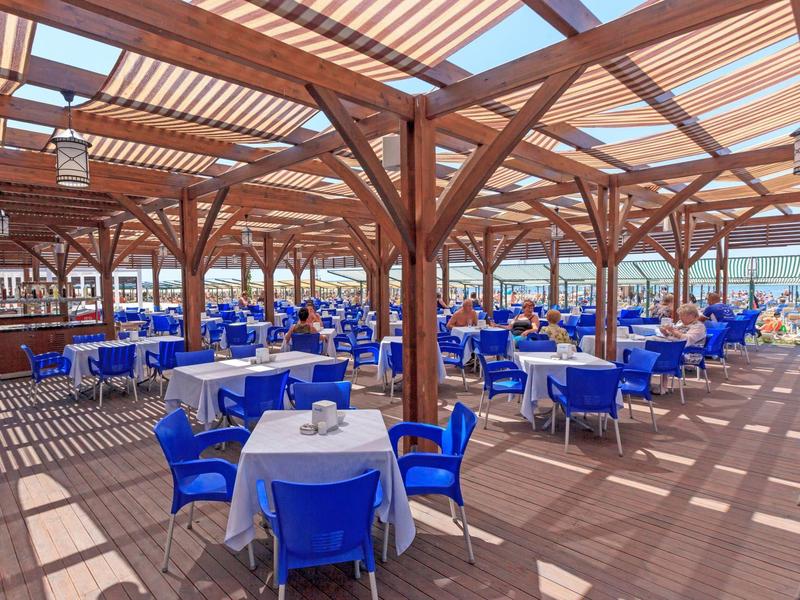 Duży zadaszony taras z niebieskimi krzesłami i białymi stołami w słonecznym otoczeniu.