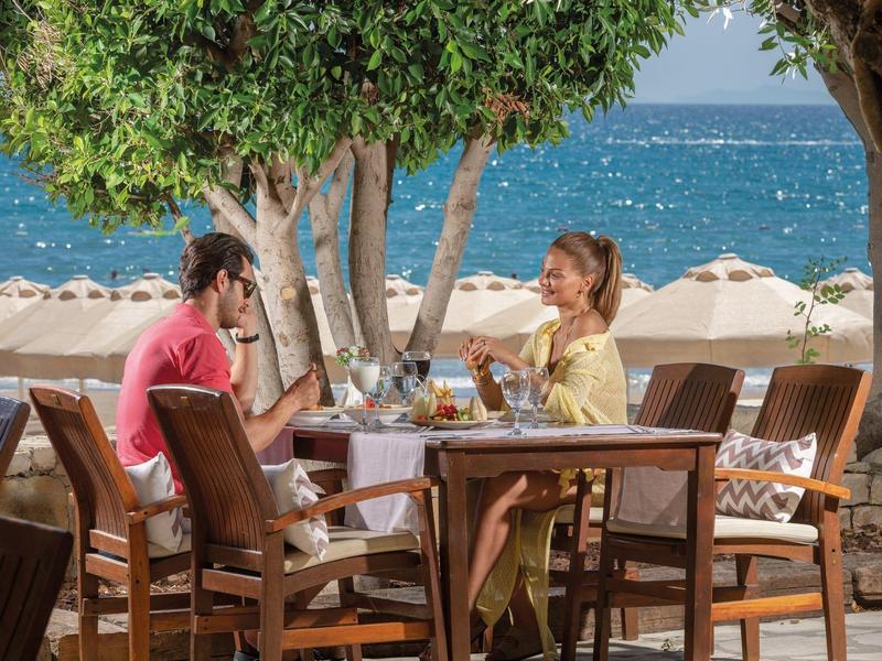 Coppia che gode di un pasto all'aperto con vista mare sotto alberi e ombrelloni.