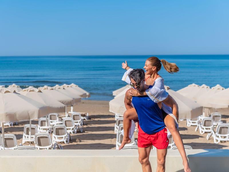 Coppia sulla spiaggia con ombrelloni e sedie, l'uomo porta la donna sulla schiena.
