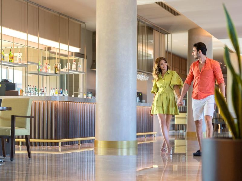 Coppia con valigie cammina in un moderno atrio dell'hotel con piante e sedie.