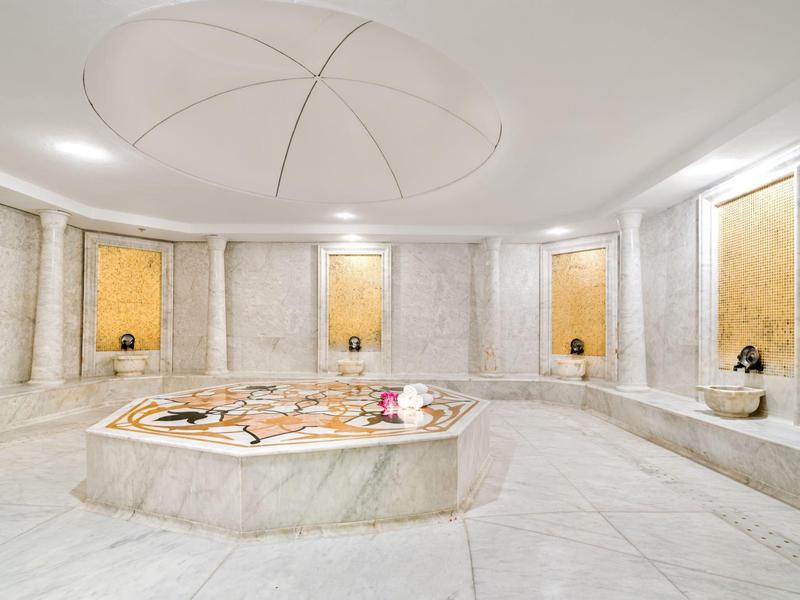 Interno luminoso in marmo bianco di un bagno turco con vasca centrale e panchine di pietra.