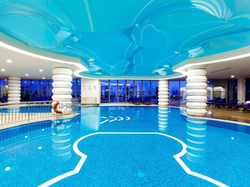 Piscina interna luminosa con acqua blu, soffitto decorato e colonne bianche in un hotel.