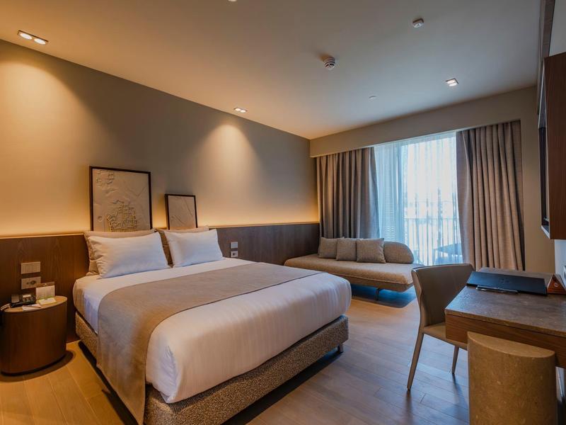 Chambre d'hôtel moderne avec grand lit, canapé et bureau près d'une grande fenêtre.