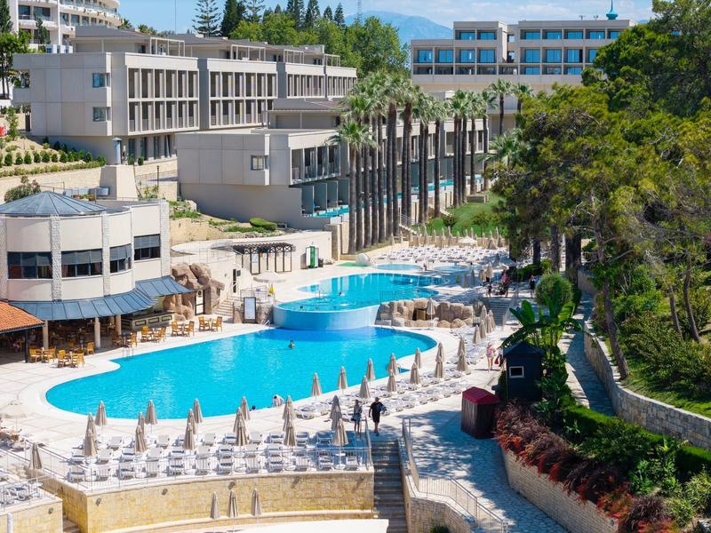 Grand piscine d'hôtel avec parasols et chaises longues, entourée de bâtiments modernes et de verdure.