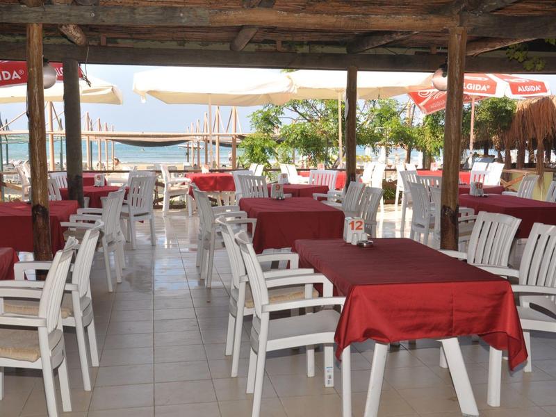 Área exterior cubierta de un restaurante con sillas blancas y mesas con manteles rojos cerca de la playa.