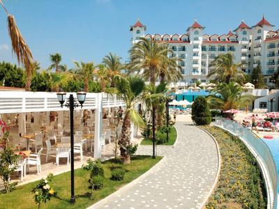 Wybrukowana ścieżka z palmami prowadząca do hotelu z basenem pod jasnym niebem.