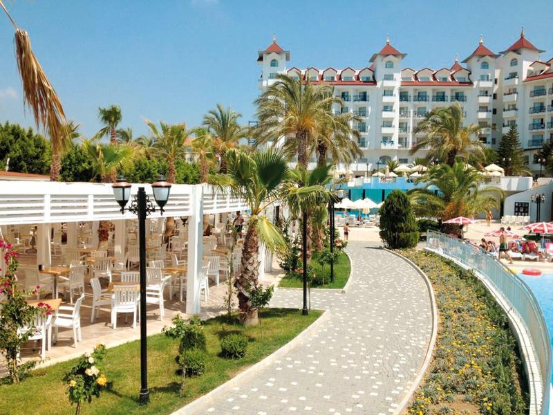 Wybrukowana ścieżka z palmami prowadząca do hotelu z basenem pod jasnym niebem.