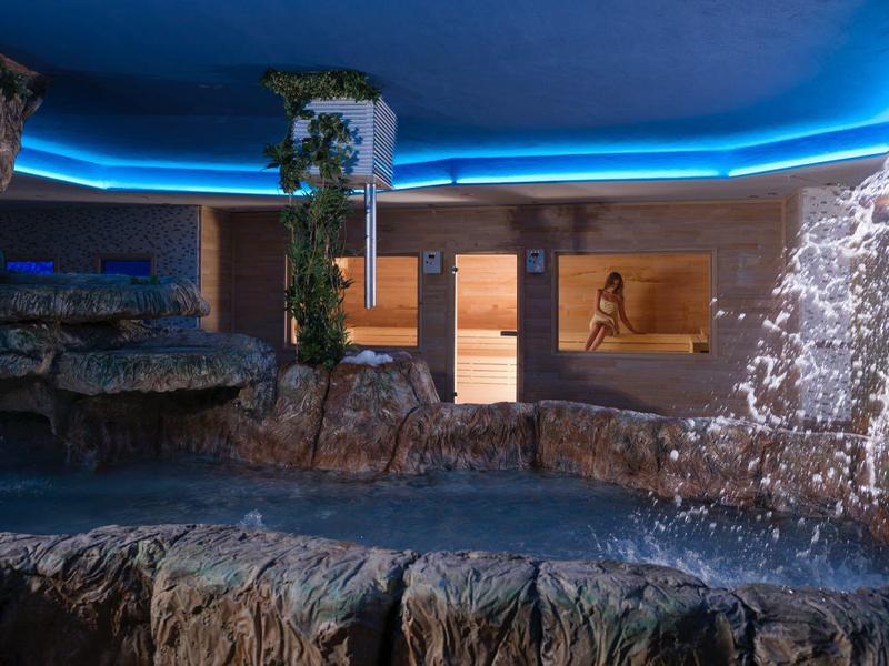 Piscina interior con paisaje rocoso, cascada y iluminación LED azul en el techo.