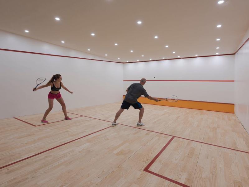 Dos personas juegan squash en una cancha interior luminosa con suelo de madera y paredes blancas.