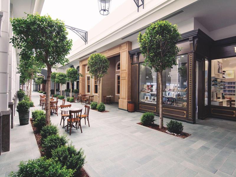 Zona peatonal cubierta con árboles, asientos y tiendas en una concurrida área comercial.