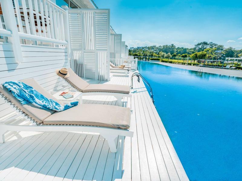 Tumbonas blancas con cojines azules en una terraza de madera junto a agua tranquila bajo cielo despejado.