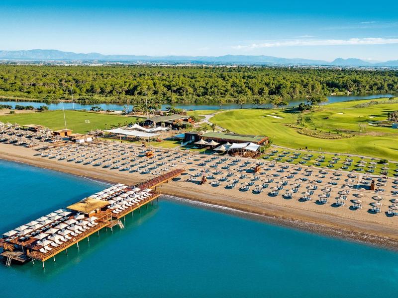Vista de una amplia playa con sombrillas, tumbonas y un muelle sobre el mar azul.