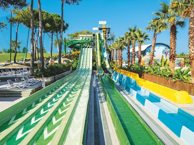 Varias toboganes de agua coloridos en un parque acuático con palmeras y tumbonas al fondo.