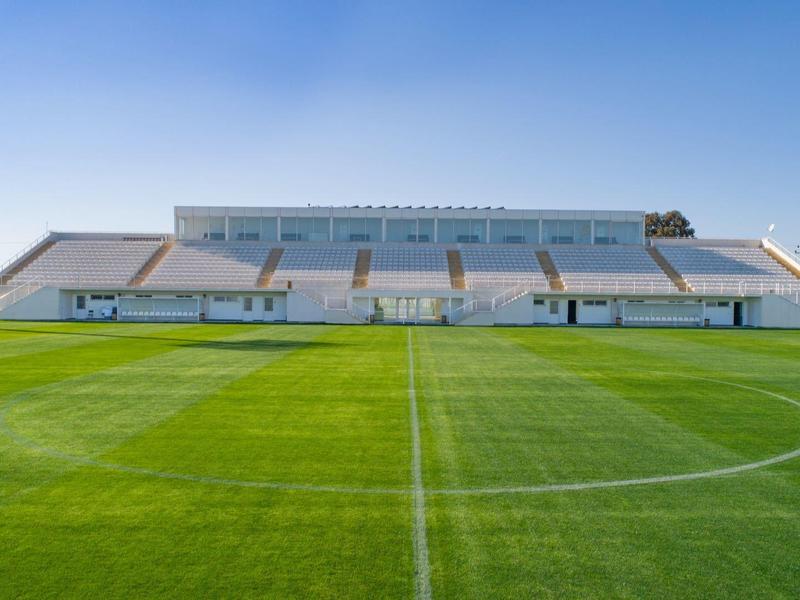 Campo de fútbol bien cuidado frente a graderías bajo un cielo despejado y azul.