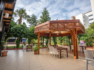 Prostorná terasa s dřevěným pavilonem, stoly a židlemi, obklopená stromy a budovami.