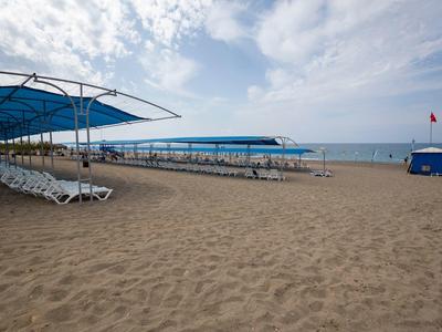Breiter Sandstrand mit blauen Sonnensegeln und weißen Liegestühlen unter bewölktem Himmel.