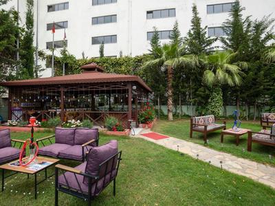 Zahradní prostor s lavičkami a pavilonem, obklopený zelenými rostlinami před bílou budovou.