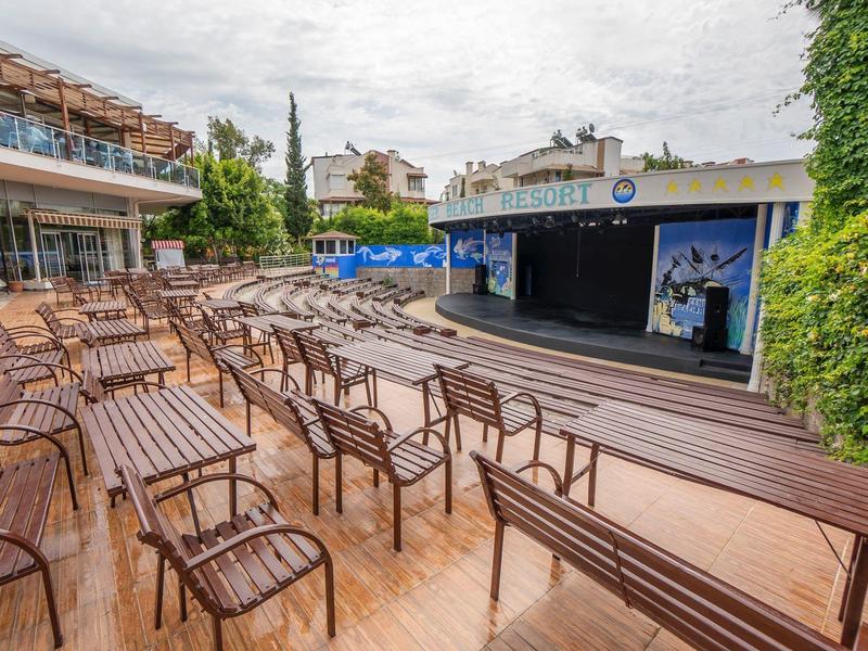 Prázdná venkovní plocha s dřevěnými lavicemi a židlemi před malým jevištěm vedle hotelu.