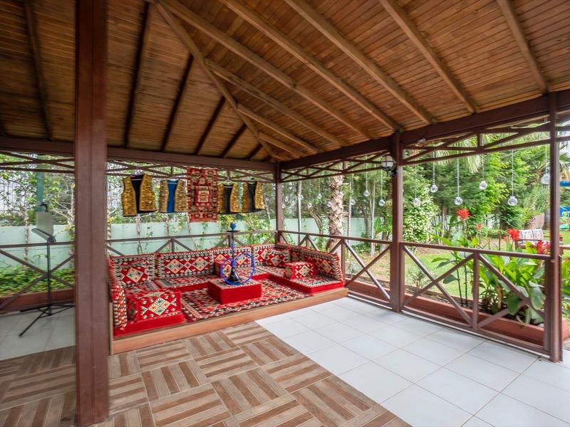 Otevřený pavilon s dřevěnou střechou, barevnými polštáři a výhledem na zahradu s jezírkem.