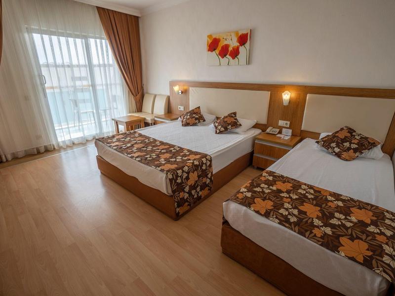 Světý hotelový pokoj se dvěma postelemi, dřevěnou podlahou a velkým oknem s závěsy.