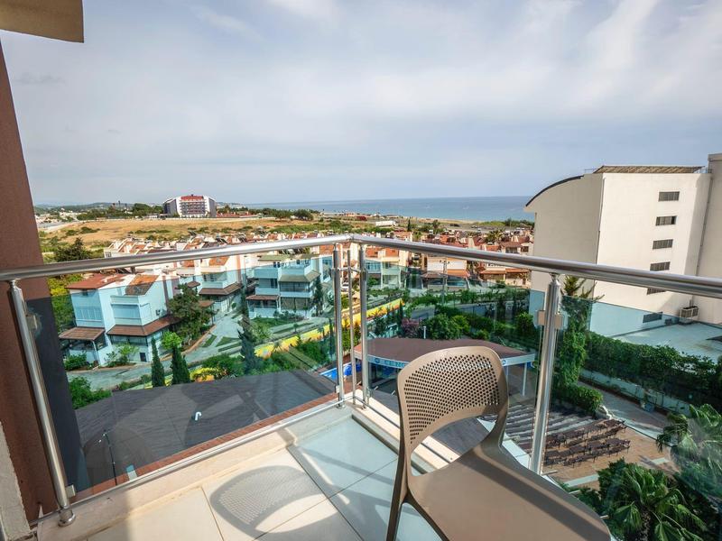 Balkon se židlí a skleněným zábradlím s výhledem na hotelové zařízení a moře v pozadí.