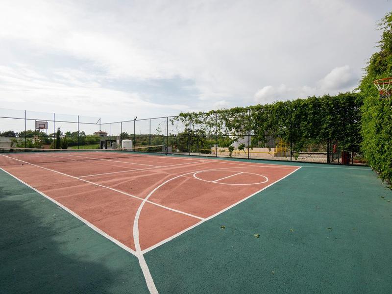 Tenisový kurt s zeleným a červeným povrchem, obklopený zelenou vegetací a částečně oblačnou oblohou.