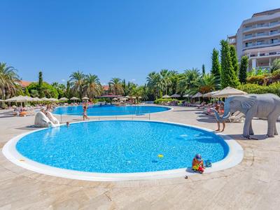 Buitenzwembad met rond kinderbad, parasols en palmbomen bij een hotel.