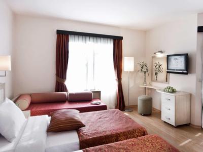 Lichte hotelkamer met tweepersoonsbed, bank, flatscreen-tv en grote ramen met gordijnen.