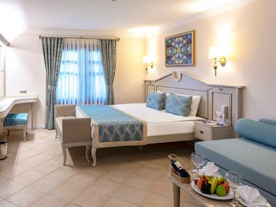 Helles Hotelzimmer mit Doppelbett, Couch, TV und einem kleinen Esstisch mit Obst.