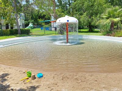 Ein flacher Wasserspielplatz mit Fontäne und Sandspielzeug am Rand unter Bäumen.