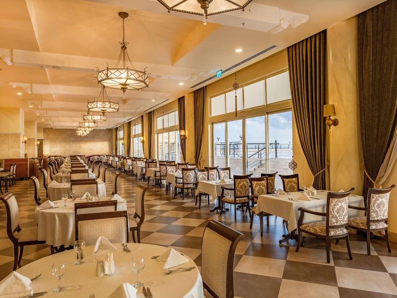 Elegante eetzaal met grote ramen, kroonluchters en comfortabele stoelen in warme kleuren.