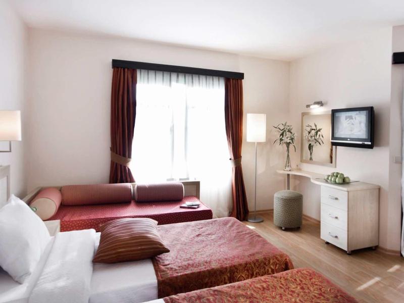 Lichte hotelkamer met tweepersoonsbed, bank, flatscreen-tv en grote ramen met gordijnen.