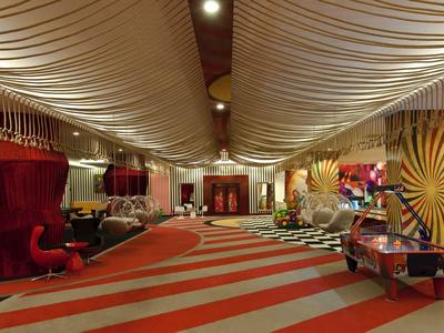 Großer, farbenfroher Hotelflur mit wellenförmiger Decke, rot-orange gestreiftem Teppich und Sitzgruppen.