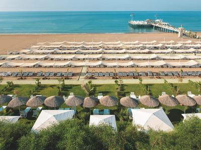 Strand mit Reihen von weißen Sonnenschirmen, Strohdächern, grüner Vegetation und Holzsteg im Meer.