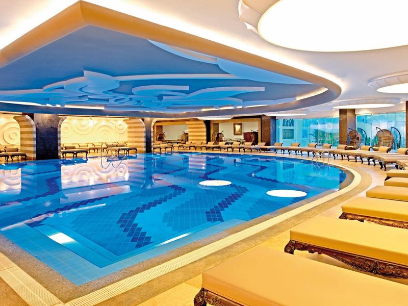 Vnitřní hotelový bazén s lehátky, modrou vodou a dekorativním stropem.