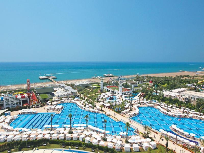 Velké letovisko s několika modrými bazény a lehátky u moře pod jasnou oblohou.