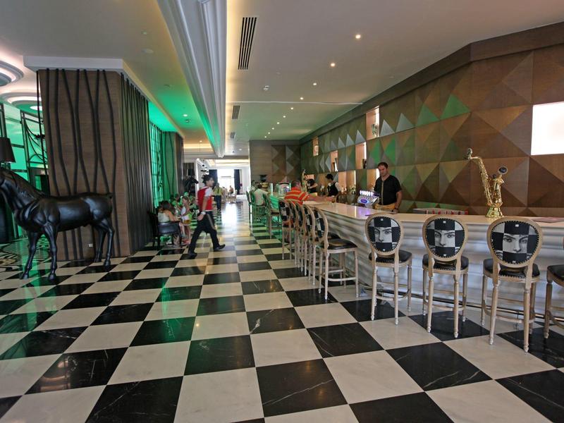 Moderní hotelový bar s černo-bílou šachovnicovou podlahou a zeleným ambientním osvětlením.