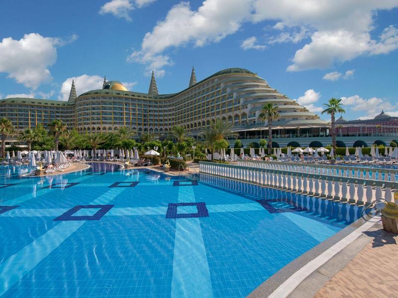 Velký moderní hotel s zakřiveným tvarem vedle prostorného venkovního bazénu pod modrou oblohou.