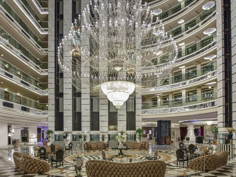Hotelová hala s velkým křišťálovým lustrem, vzorovanou podlahou a kruhovým uspořádáním sezení.