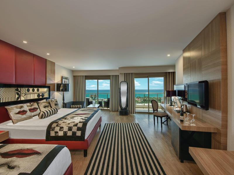 Prostorný hotelový pokoj se dvěma lůžky, pruhovaným kobercem, psacím stolem a velkými okny s výhledem na moře.