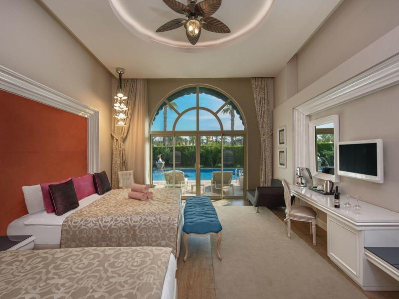 Pohodlný hotelový pokoj se dvěma postelemi, modrými stoličkami a velkým oknem s výhledem na palmy a bazén.