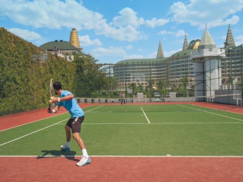 Muž hraje tenis na tenisovém kurtu s velkou hotelovou budovou v pozadí.